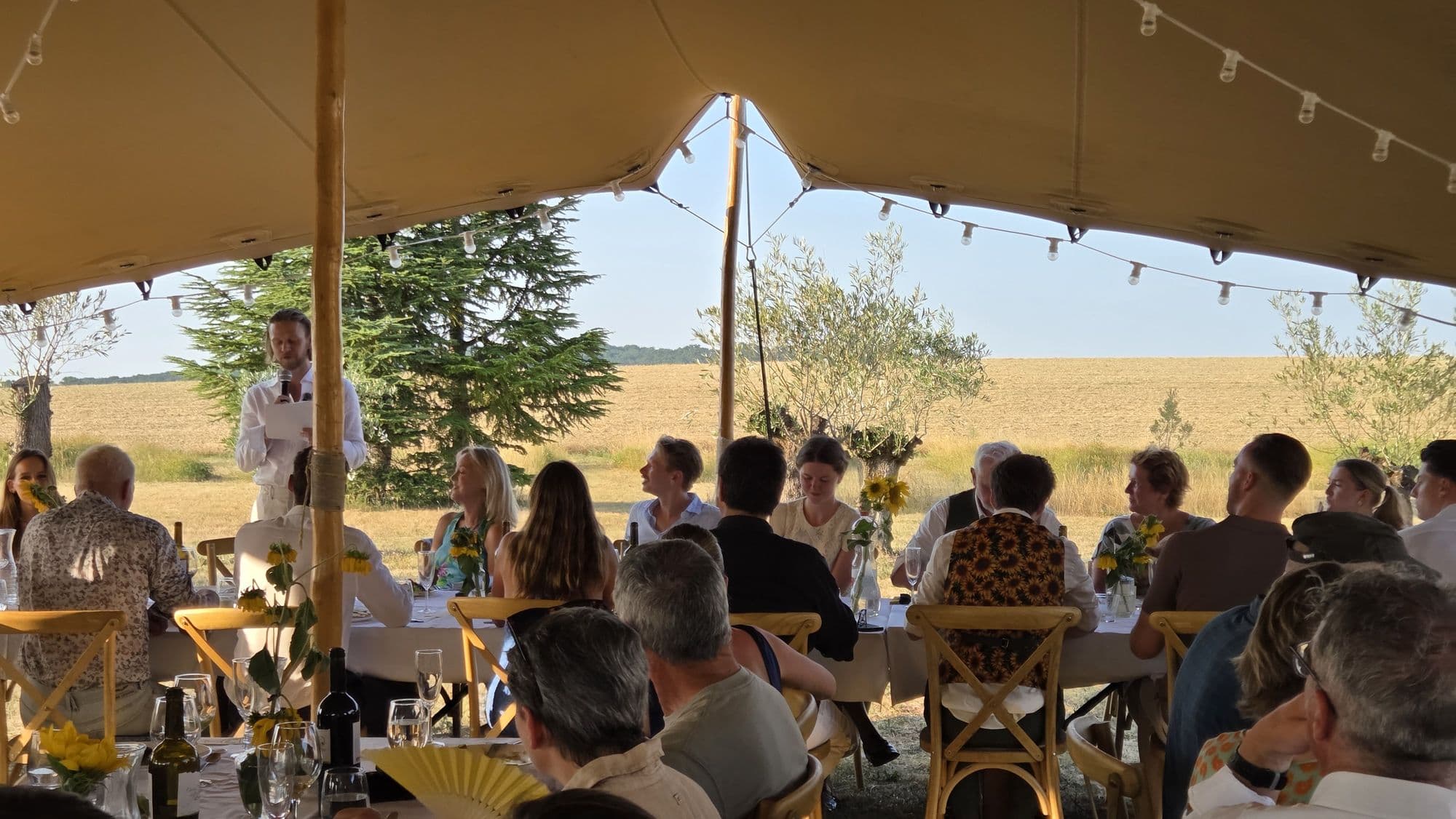 Wedding guests at long tables inside a stretch marquee, festoon lights above, fields beyond